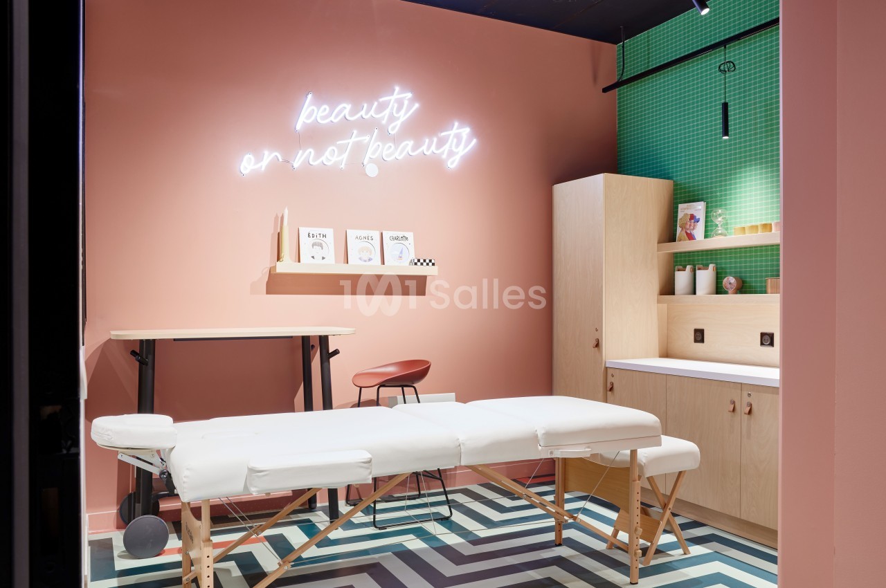 Salle moderne avec table de massage, mur rose, néon ’beauty or not beauty’ et étagères en bois sur fond vert.
