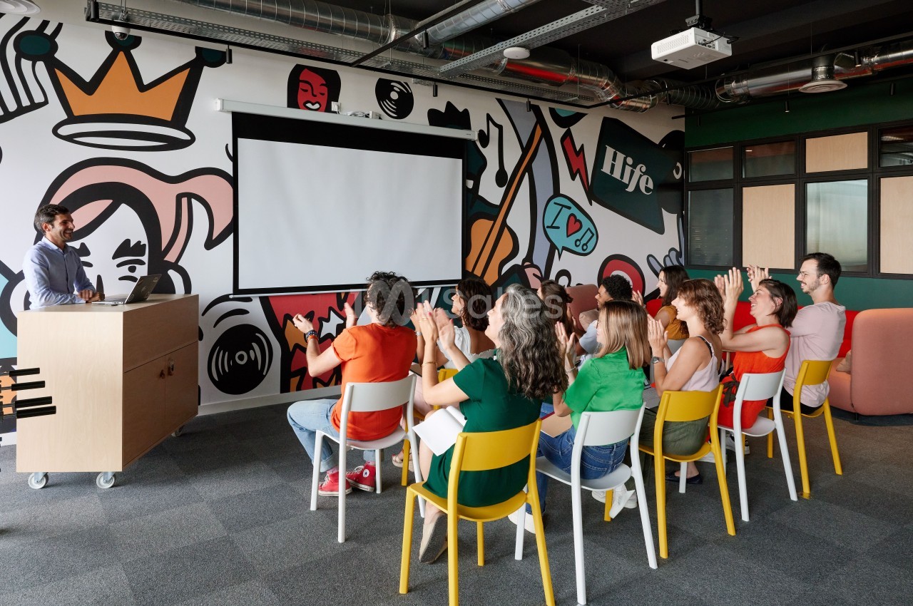 Un groupe de personnes assises dans une salle colorée écoute un présentateur devant un écran de projection.