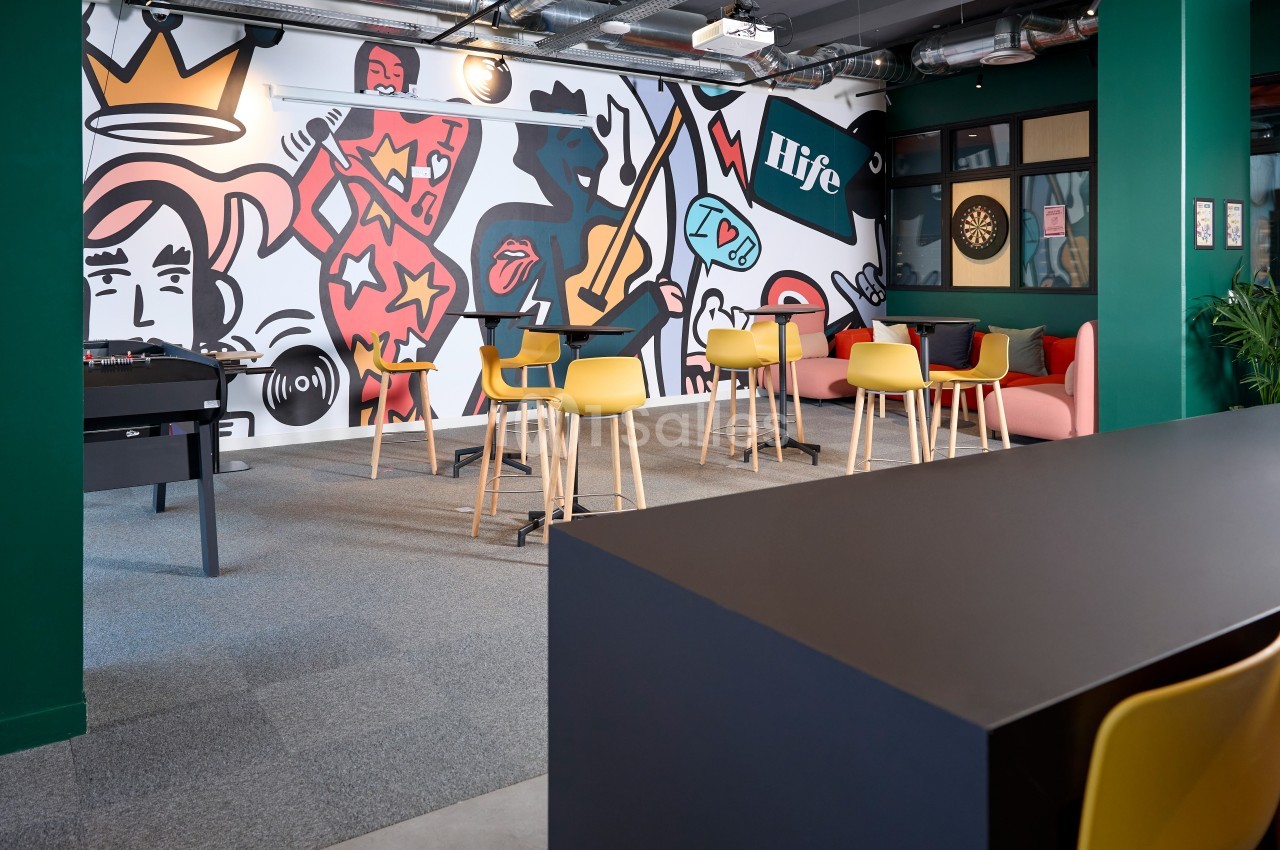 Espace de détente avec des tables hautes, des chaises jaunes, un baby-foot et une fresque murale colorée.