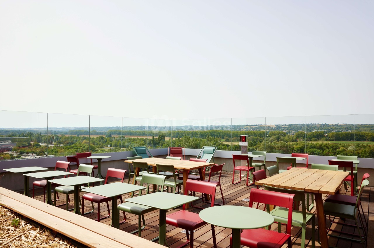 Terrasse avec tables et chaises colorées, vue dégagée sur un paysage verdoyant sous un ciel clair.