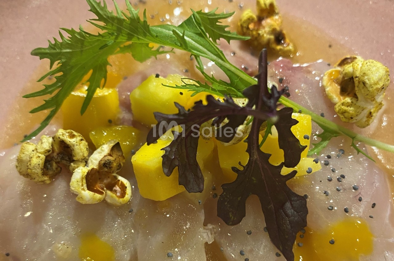 Tranches de poisson cru avec dés de mangue, feuilles de salade, maïs soufflé et graines noires sur une assiette.