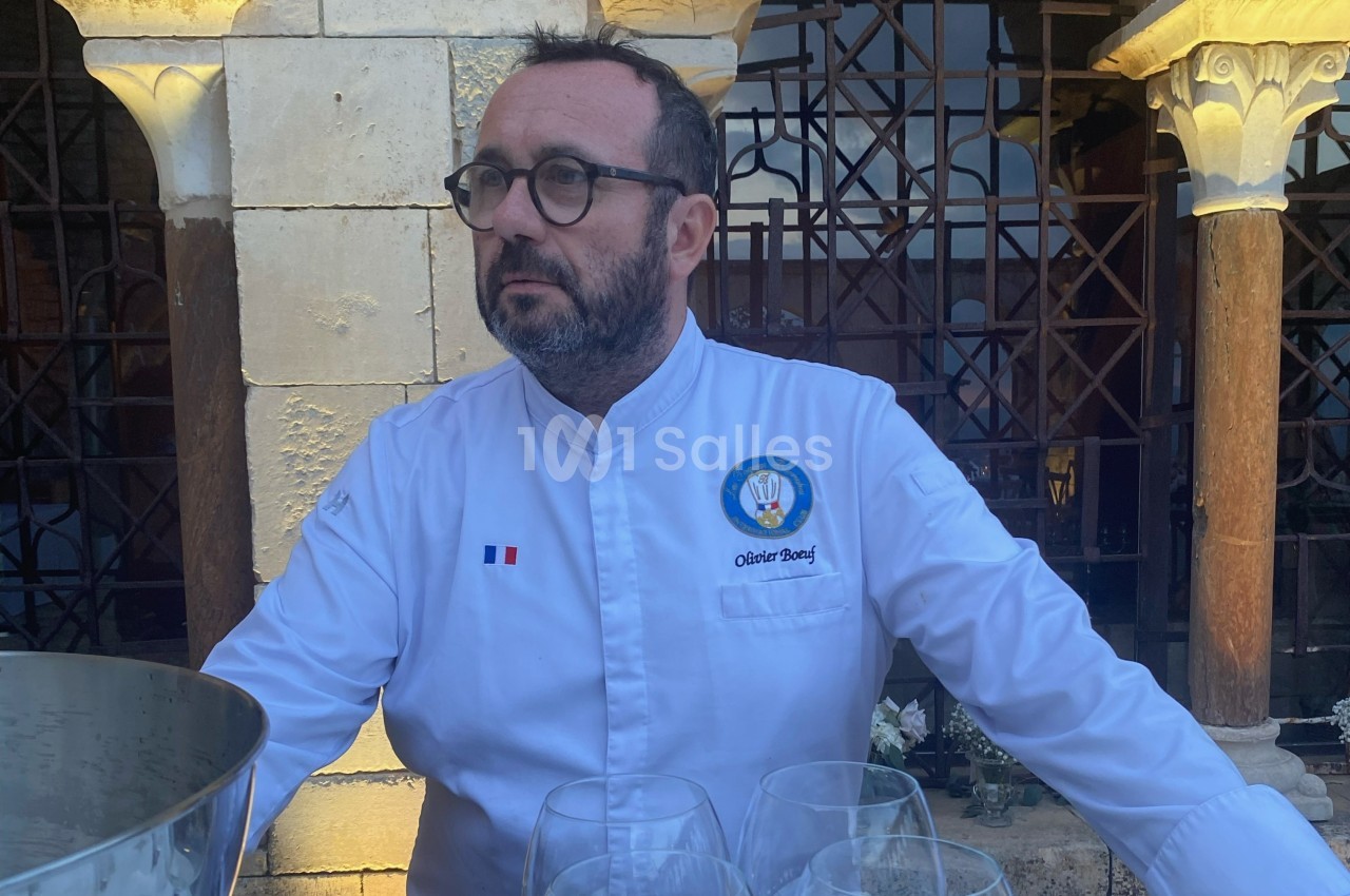 Homme en veste de chef blanche, debout devant une architecture en pierre et des grilles métalliques.
