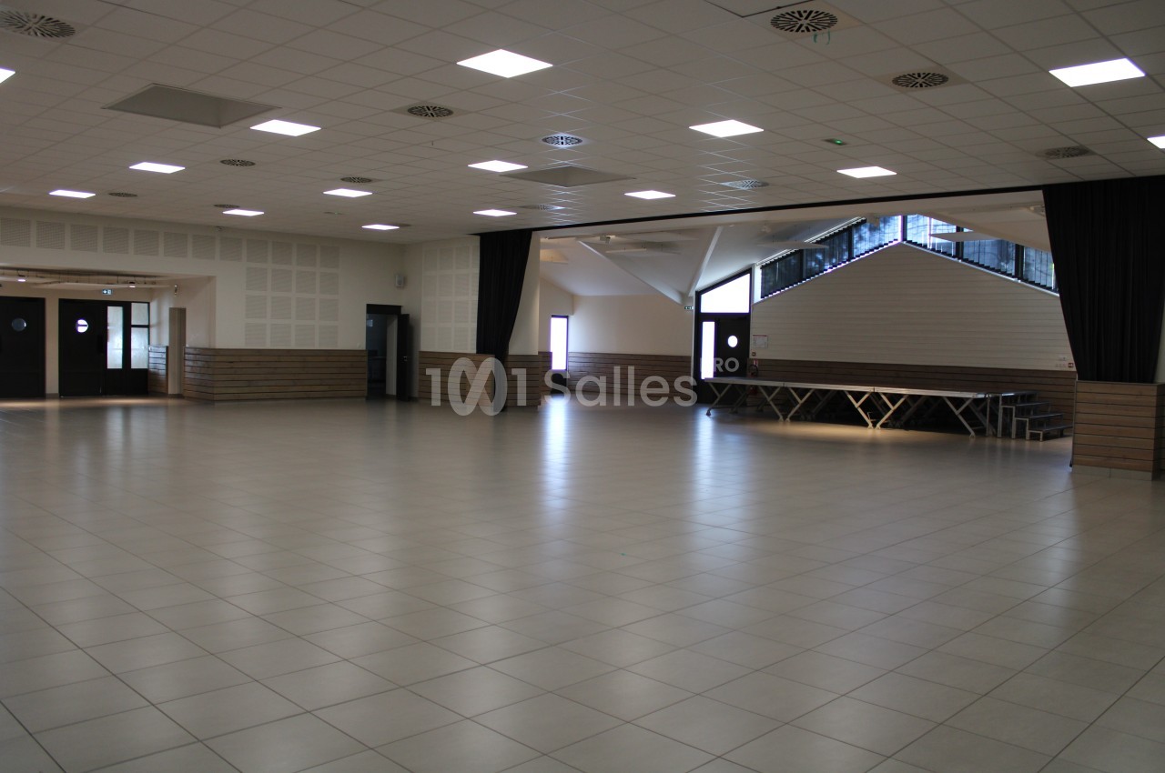 Salle spacieuse avec sol carrelé, murs clairs, plafond à dalles lumineuses et estrade en bois au fond.