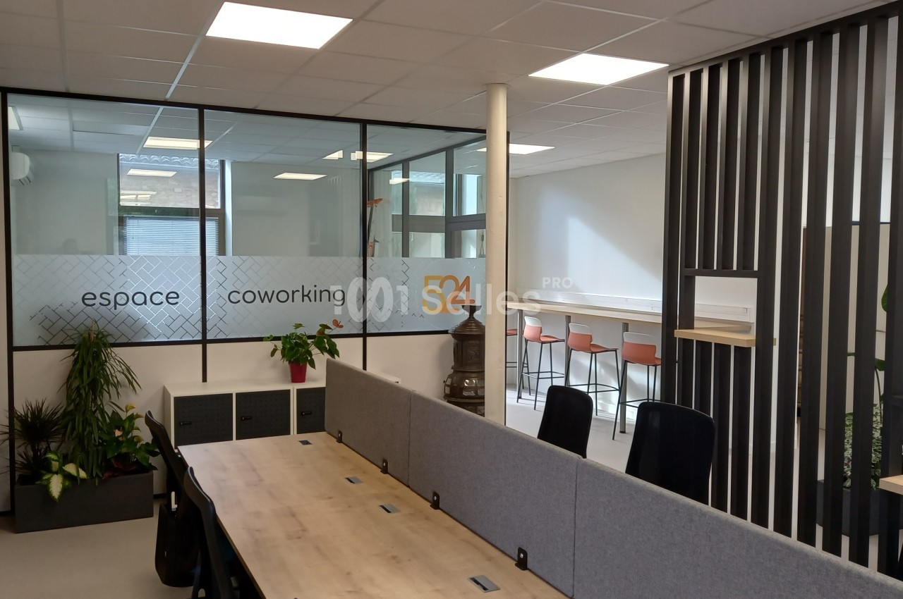 Espace de coworking lumineux avec bureaux partagés, plantes décoratives et coin café avec tabourets hauts.