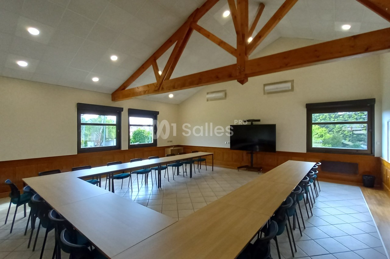 Salle de réunion lumineuse avec tables en U, chaises, écran de présentation et poutres apparentes au plafond.