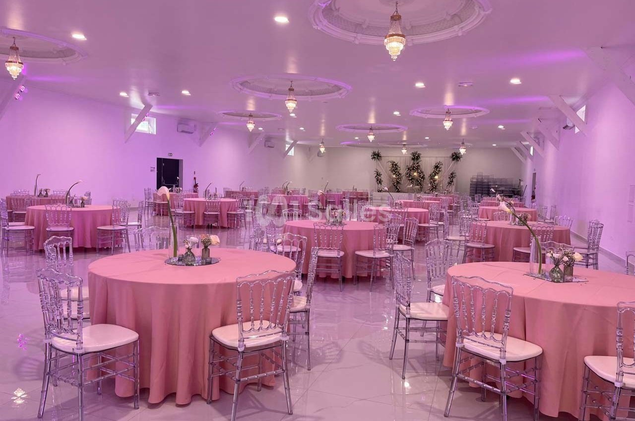 Salle de réception décorée avec des tables rondes nappées de rose, chaises transparentes et éclairage tamisé.