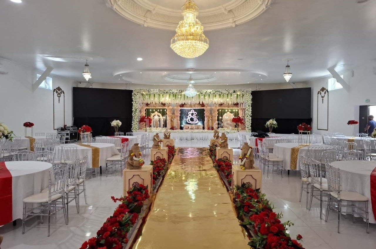 Salle de réception décorée avec des tables, des chaises transparentes, un podium fleuri et une allée dorée ornée de roses…