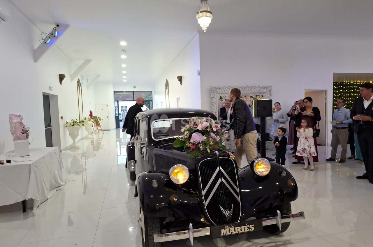 Voiture ancienne noire décorée de fleurs avec une plaque ’Mariés’, entourée d'invités dans une salle lumineuse.