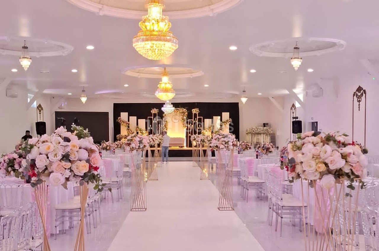 Salle de réception décorée pour un mariage avec chandeliers, allée centrale bordée de fleurs et estrade ornée.