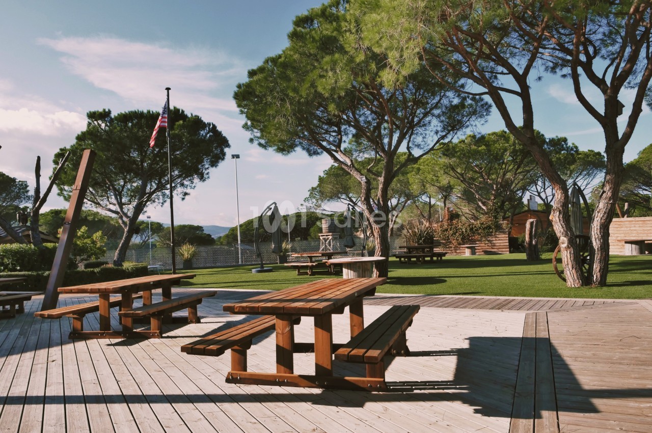 Tables de pique-nique en bois sur une terrasse ensoleillée, entourées d'arbres et d'une aire de jeux en arrière-plan.