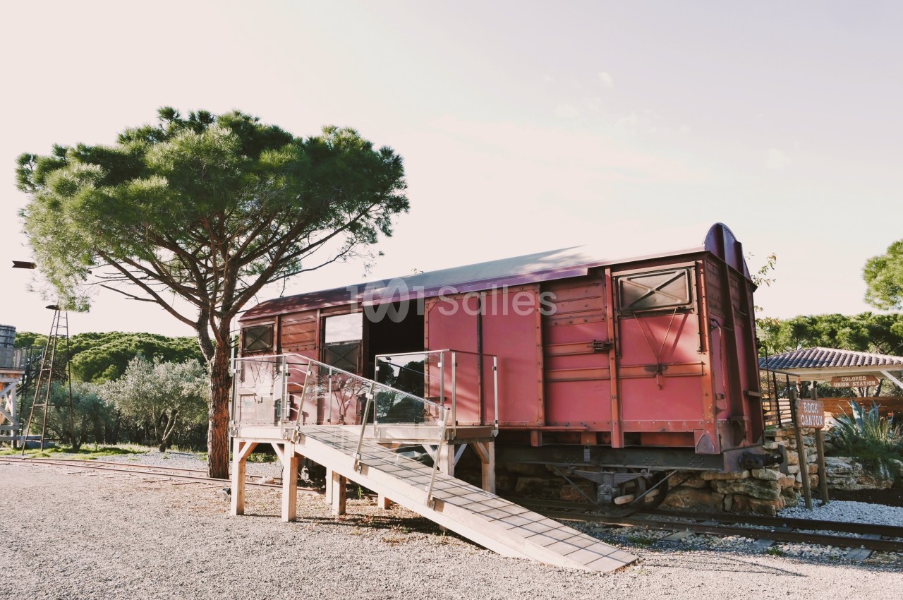 Wagon rouge aménagé avec une rampe d'accès en bois, situé près d'un arbre et entouré de végétation.