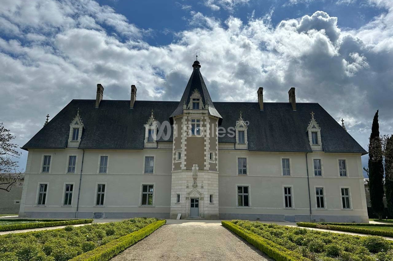 Façade d'un château à l'architecture classique, entouré de jardins symétriques sous un ciel partiellement nuageux.