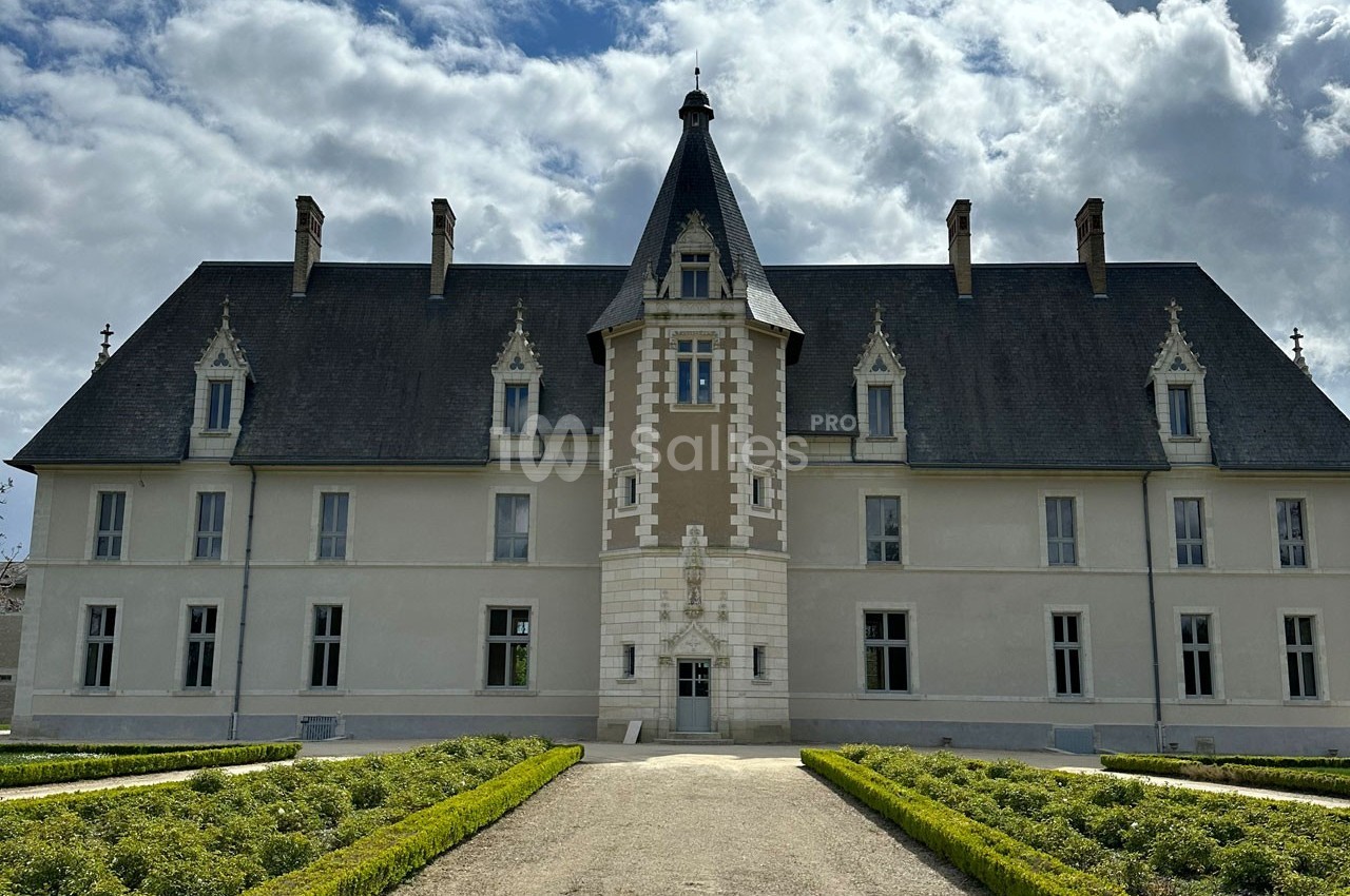 Façade d'un château avec une tour centrale, entouré de jardins symétriques sous un ciel partiellement nuageux.