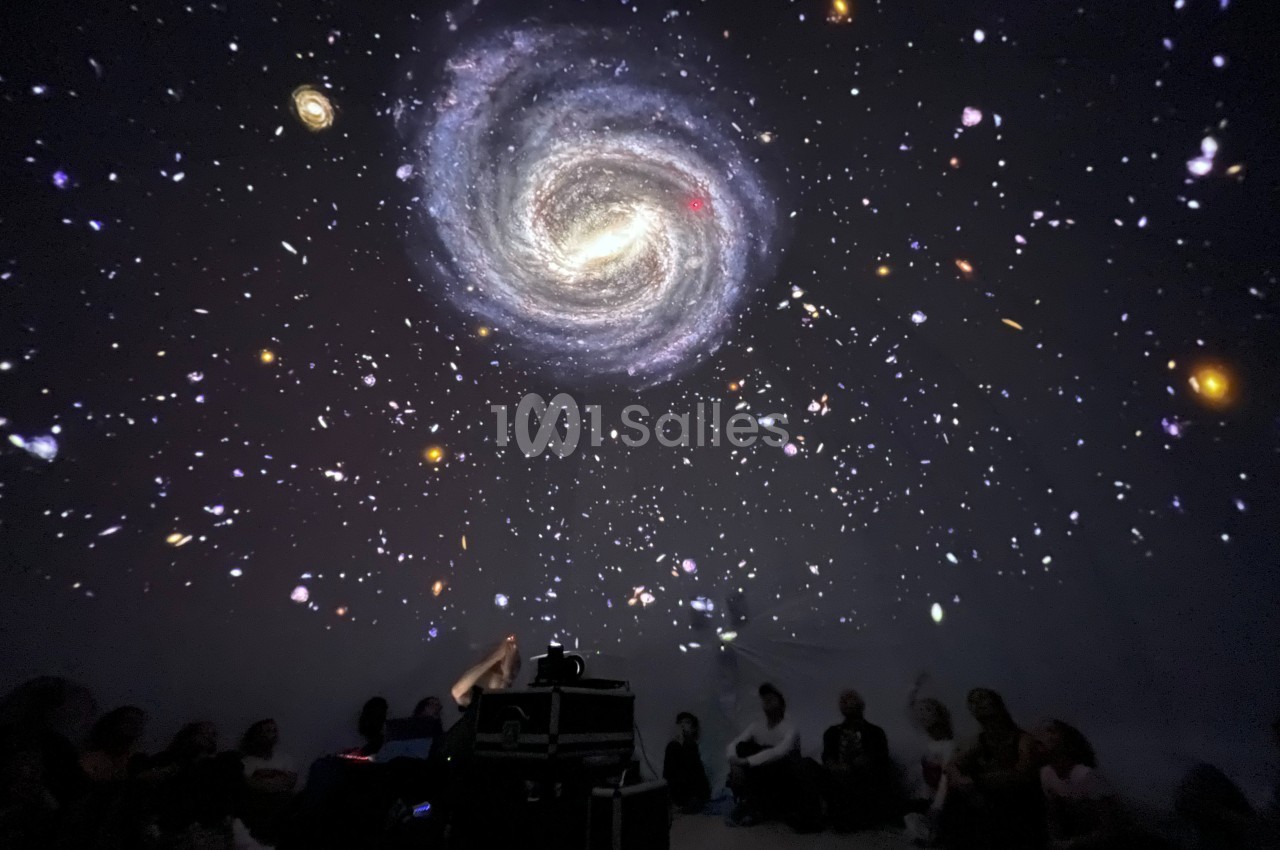 Projection immersive d'une galaxie spirale et d'étoiles dans une salle sombre, avec des spectateurs assis au sol.