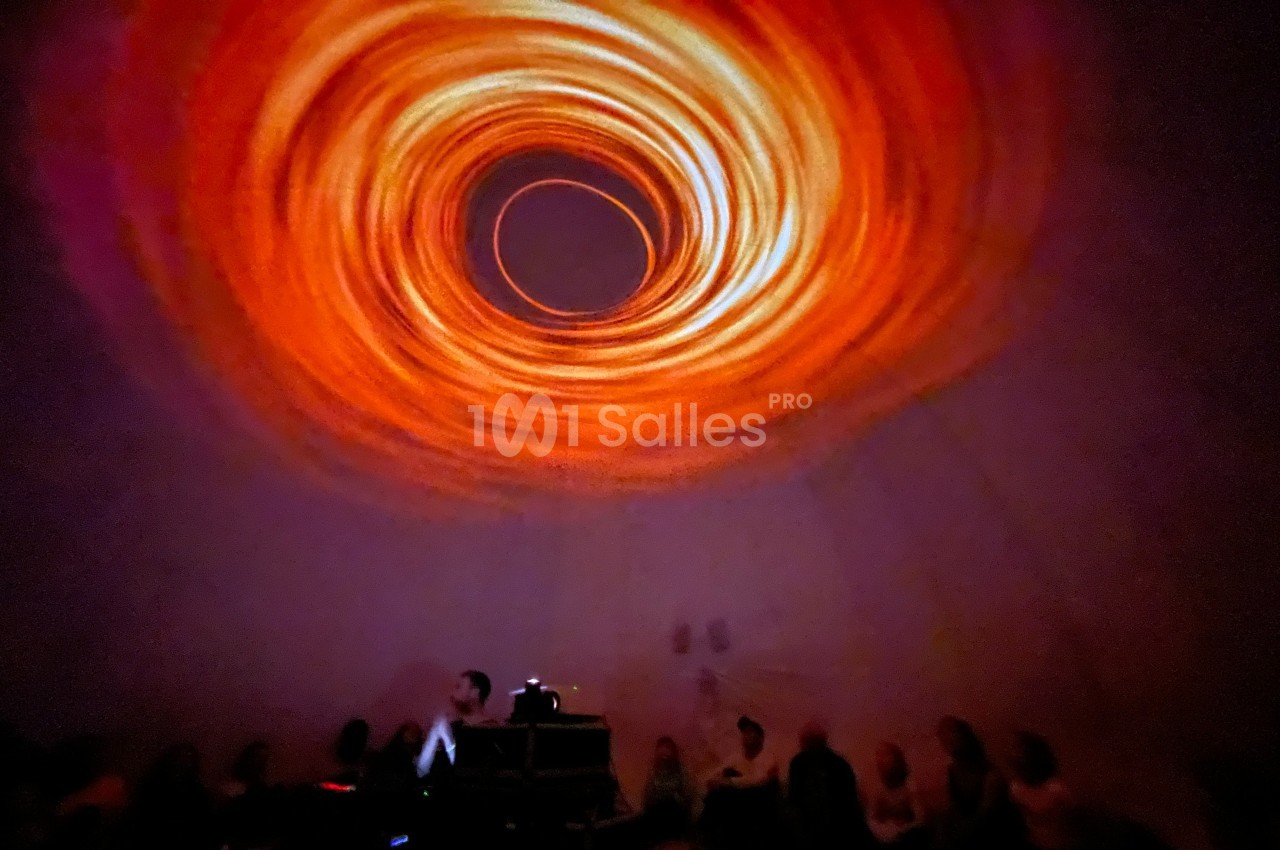 Projection lumineuse en spirale orange sur un plafond, avec des silhouettes de spectateurs assis en contrebas.