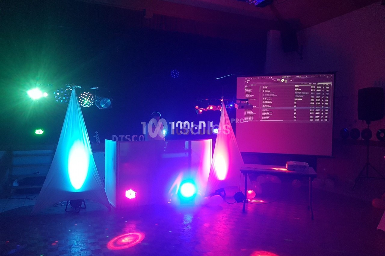 Un DJ mixe sur une scène éclairée par des lumières colorées dans une salle sombre avec un écran projeté.