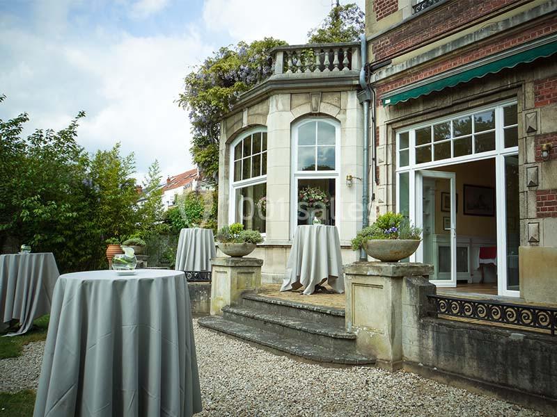 Terrasse d'une maison en pierre avec des tables hautes drapées de nappes grises, entourée de verdure.