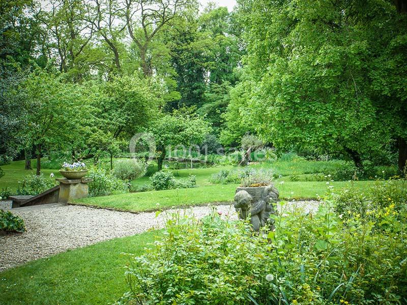 Jardin verdoyant avec pelouse, parterres fleuris, statue en pierre et arbres entourant un petit étang.