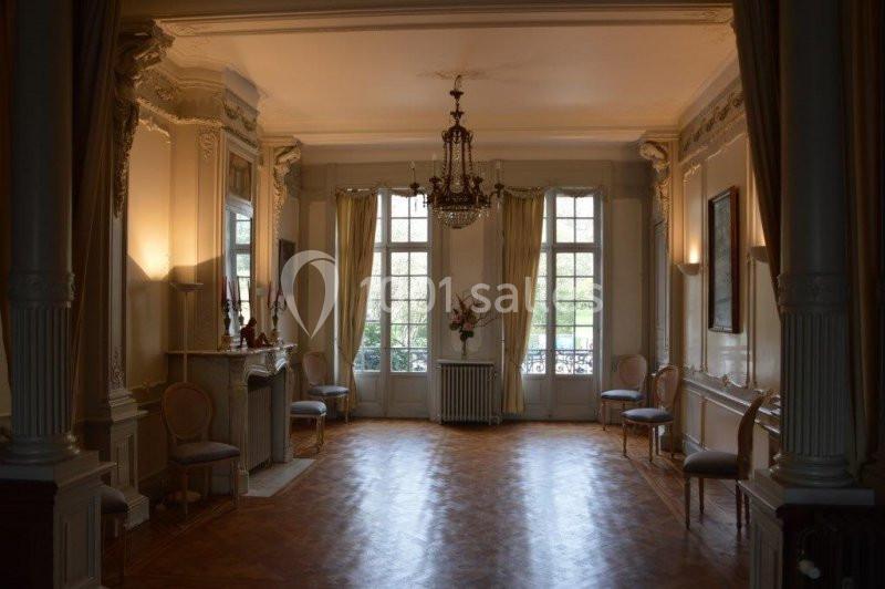 Salle élégante avec parquet, moulures, lustre, grandes fenêtres et chaises alignées le long des murs.