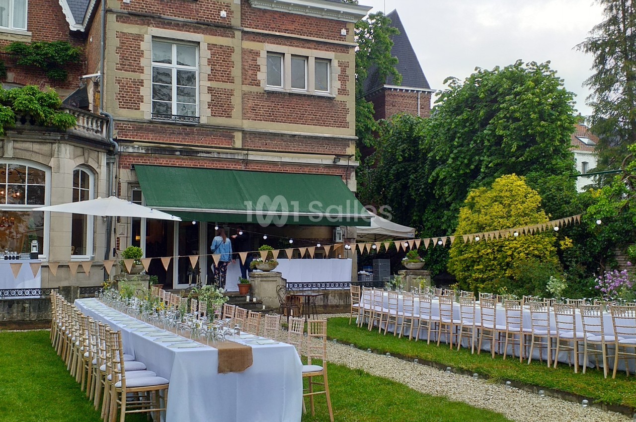 Tables dressées pour un événement en plein air dans un jardin, devant une maison en briques avec des guirlandes décoratives.