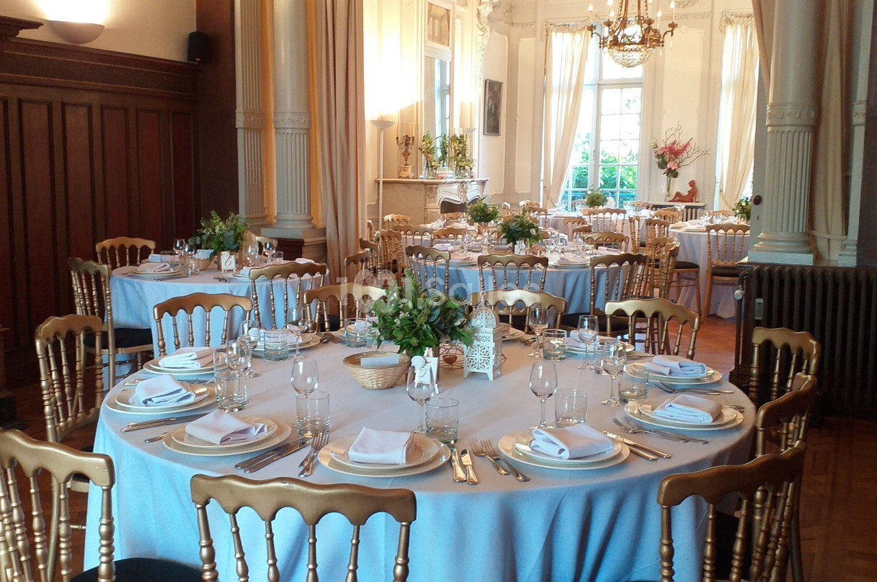 Salle élégante avec tables rondes dressées, nappes bleues, chaises dorées et lumière naturelle entrant par de grandes…