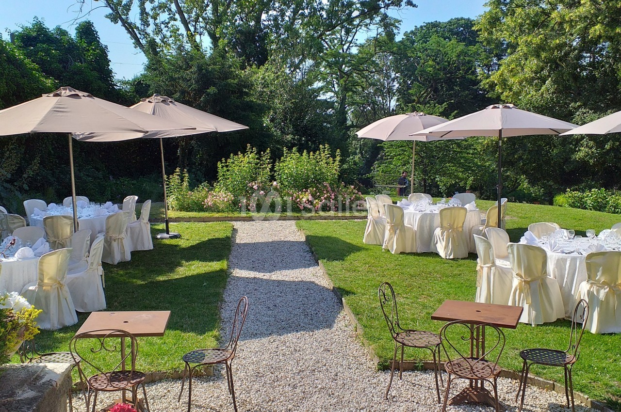 Tables dressées avec nappes blanches et chaises, disposées dans un jardin verdoyant sous des parasols beiges.