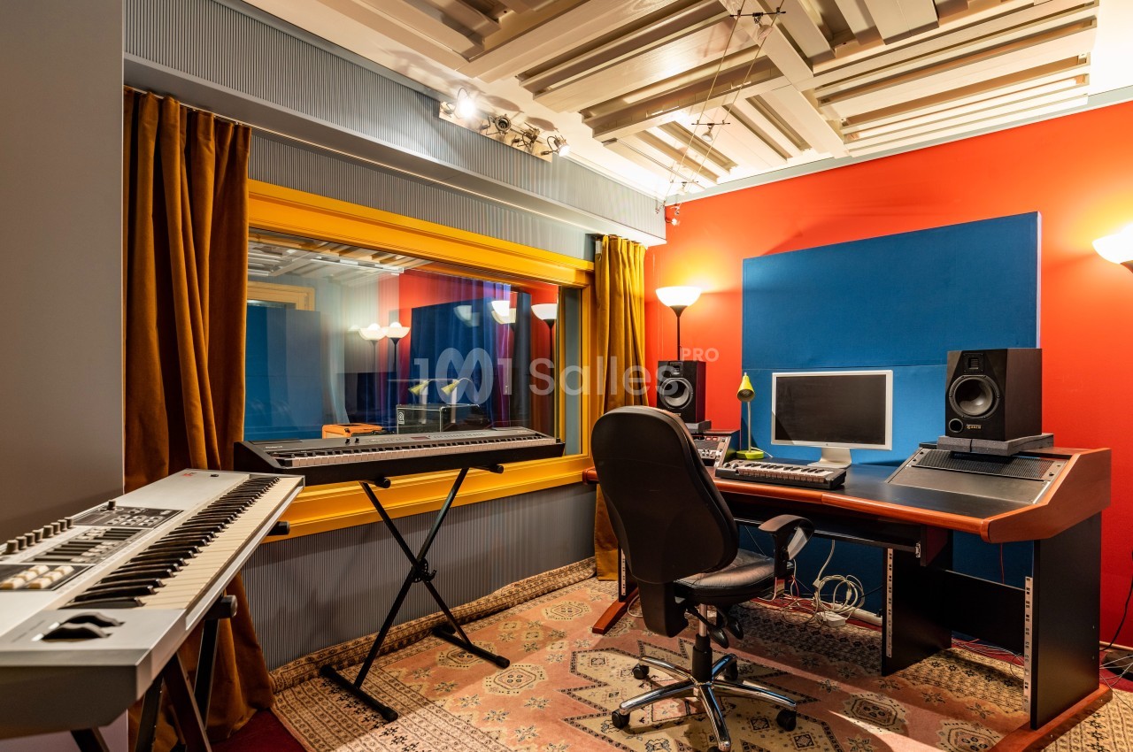 Studio d'enregistrement avec clavier, bureau équipé d'un écran et d'enceintes, et cabine insonorisée vitrée.