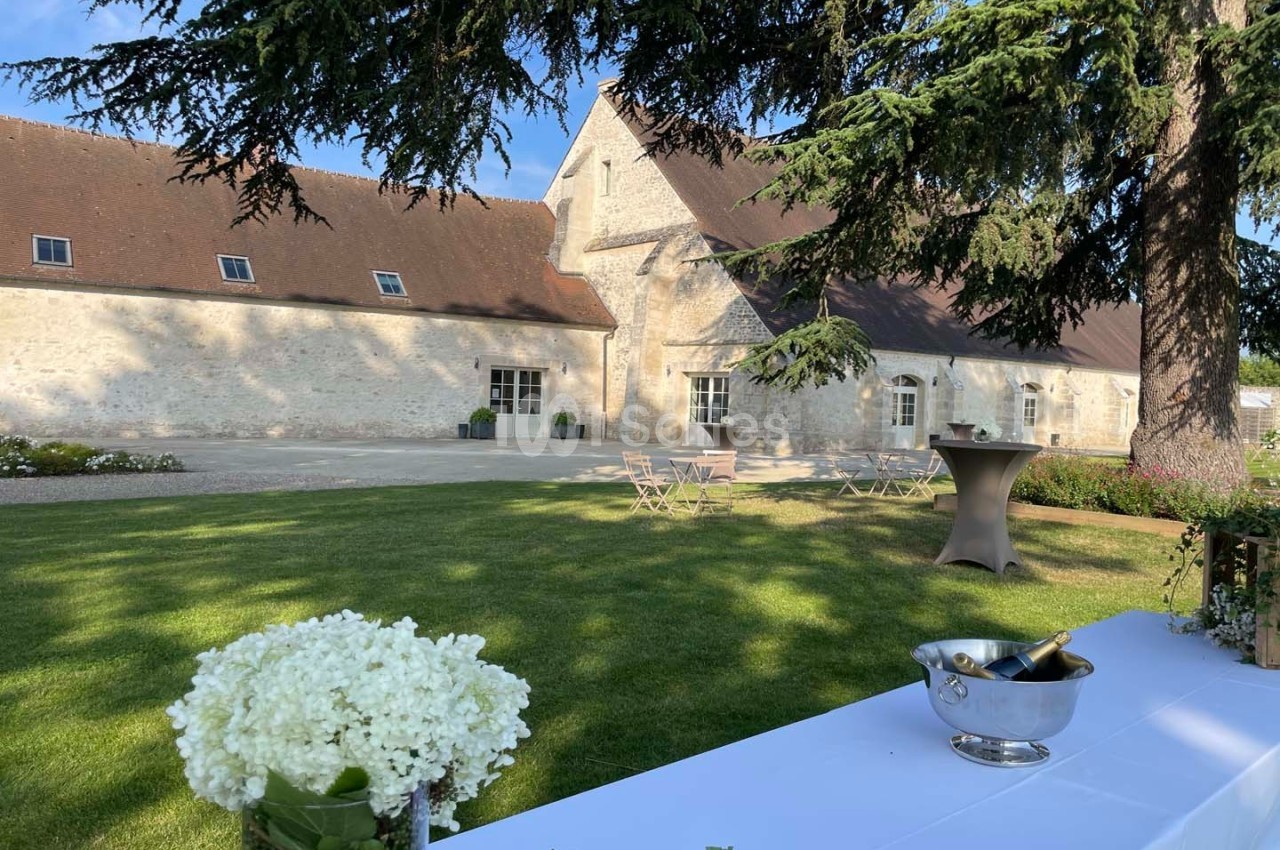 Cour d'une bâtisse en pierre avec pelouse, tables de jardin, et une table décorée de fleurs blanches au premier plan.