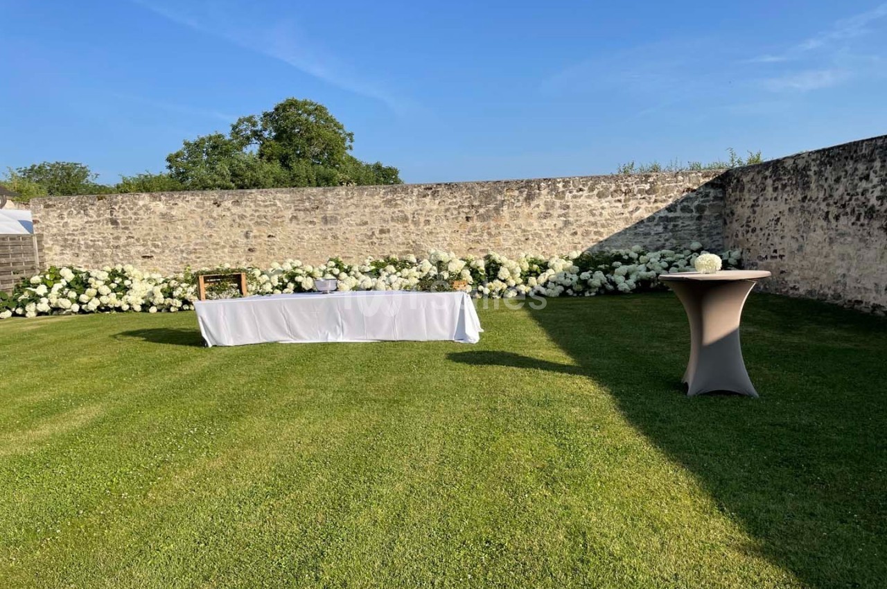 Pelouse avec table drapée blanche et table haute, bordées de fleurs blanches devant un mur en pierre sous un ciel dégagé.