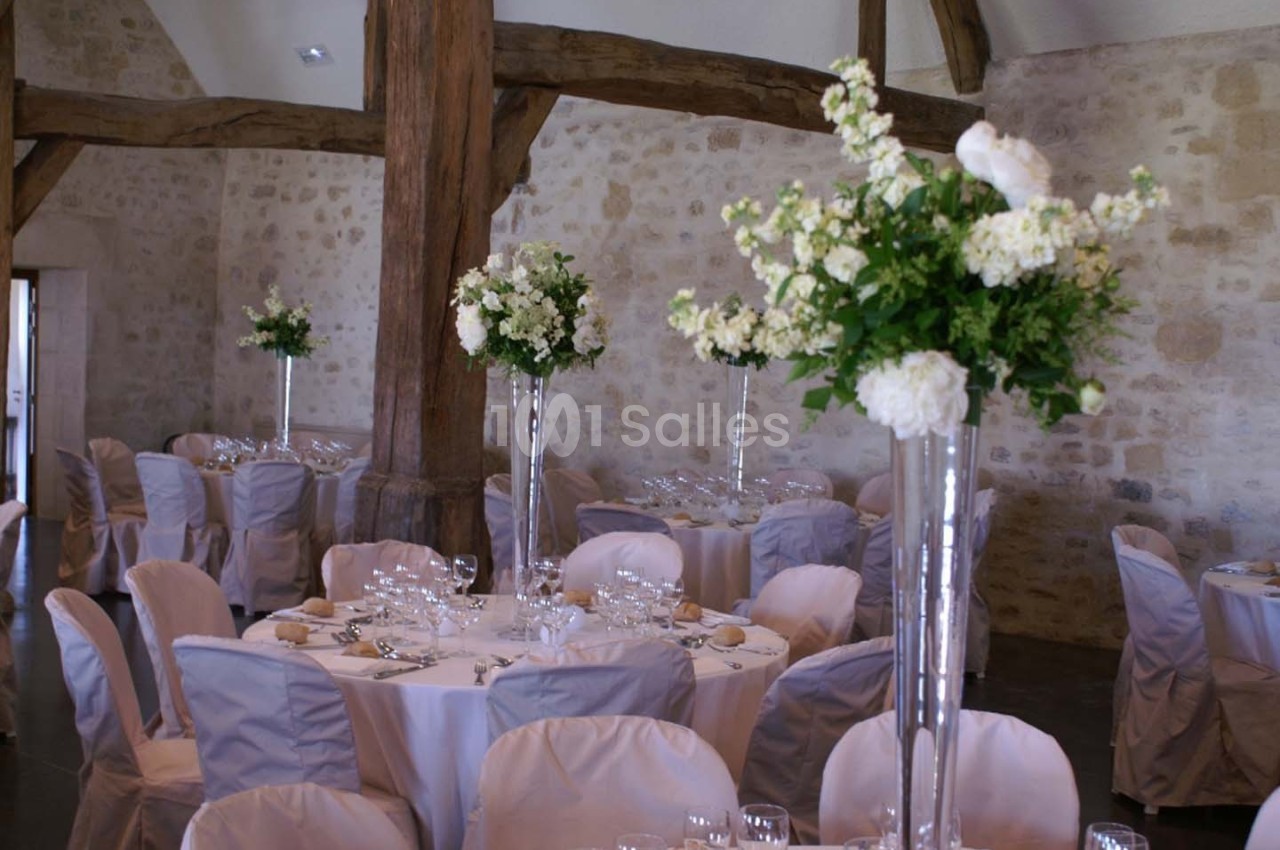 Salle de réception avec tables rondes dressées, nappes blanches et centres de table fleuris dans des vases hauts.