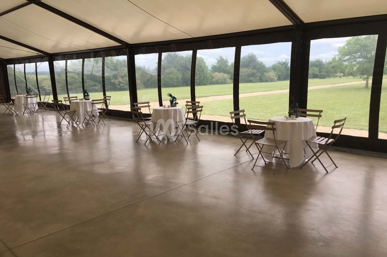 Salle avec de grandes baies vitrées donnant sur un espace vert, tables rondes dressées avec chaises en bois pliantes.