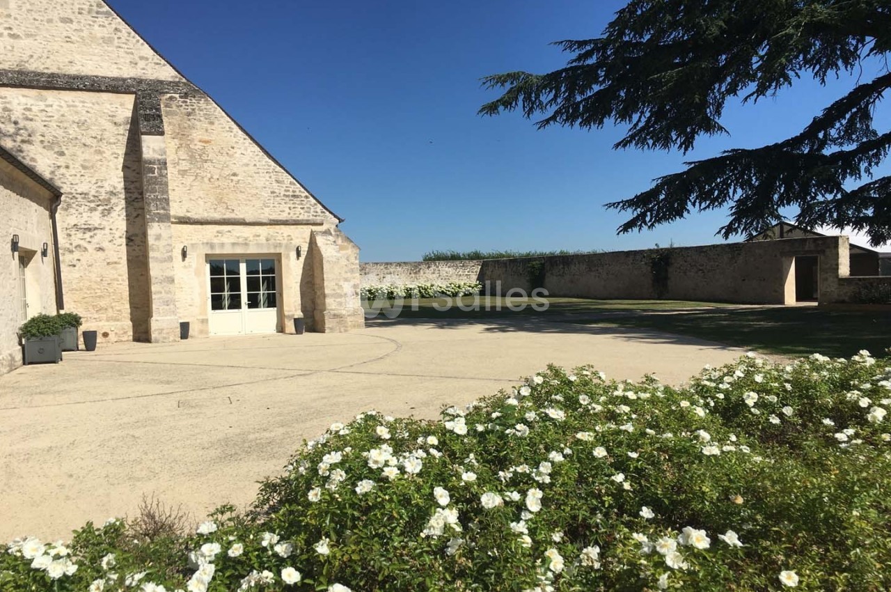 Cour ensoleillée avec façade en pierre, parterre de fleurs blanches au premier plan et mur en pierre en arrière-plan.