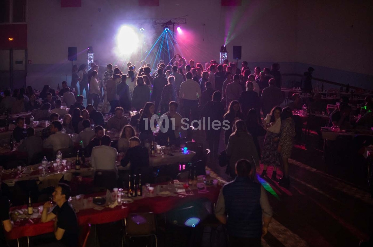 Salle remplie de convives assis et debout, éclairée par des lumières colorées et une ambiance festive.