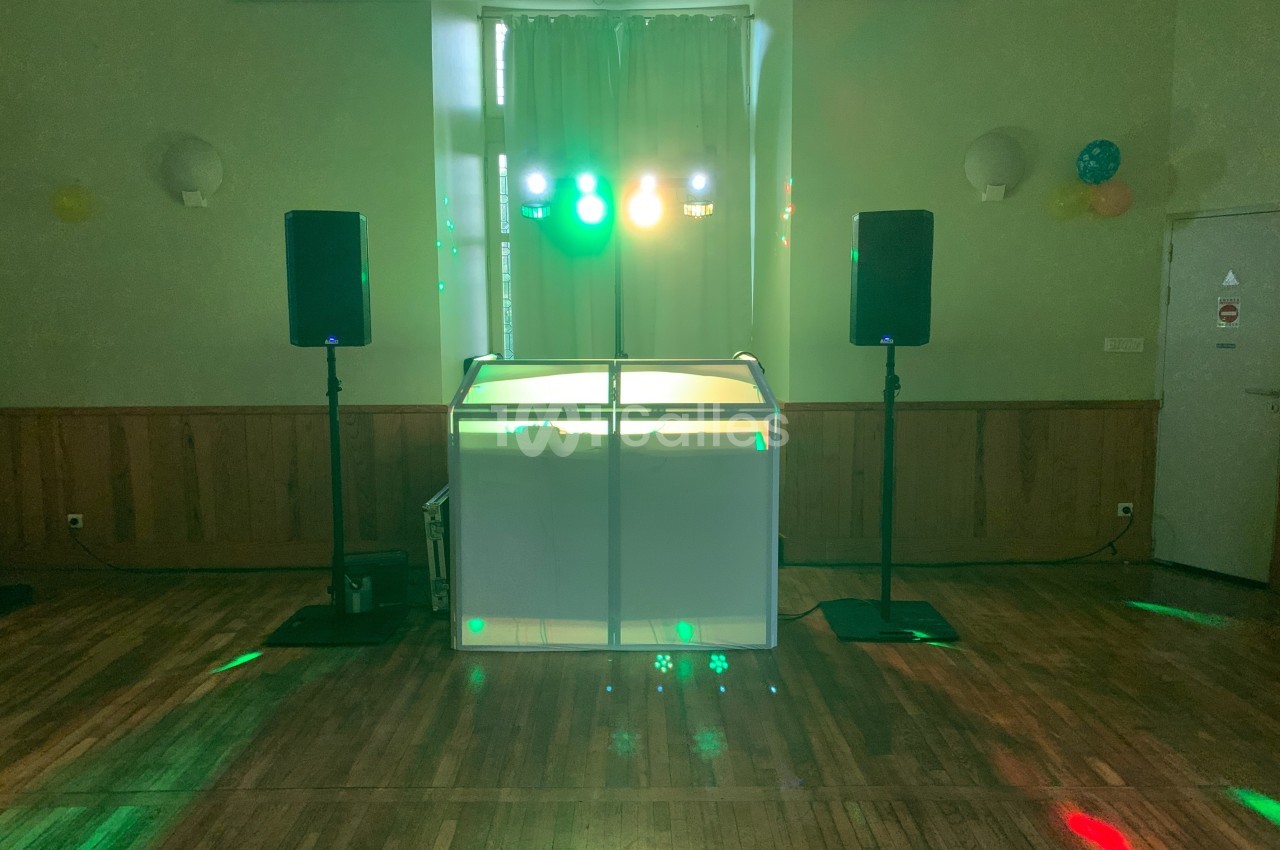 Console de DJ éclairée avec des lumières colorées, entourée de deux enceintes dans une salle au parquet en bois.