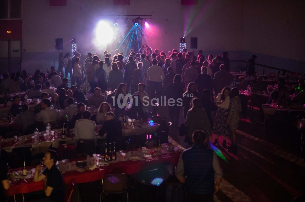 Salle remplie de convives assis et debout, éclairée par des lumières colorées et une ambiance festive.