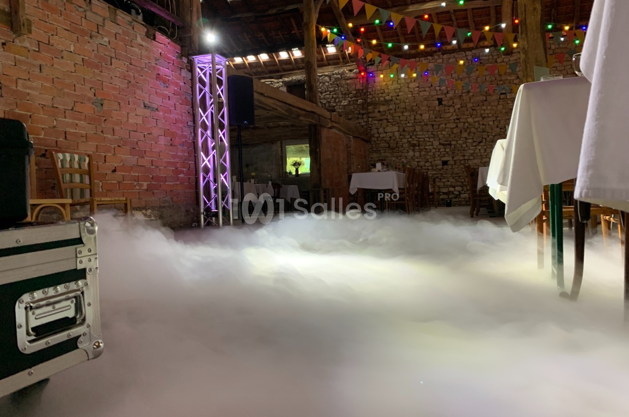 Salle en briques décorée pour un événement, avec tables dressées, guirlandes lumineuses et sol recouvert de fumée…