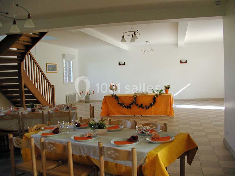 Salle lumineuse avec tables dressées pour un repas, nappes jaunes et oranges, décorations simples et escalier en bois.