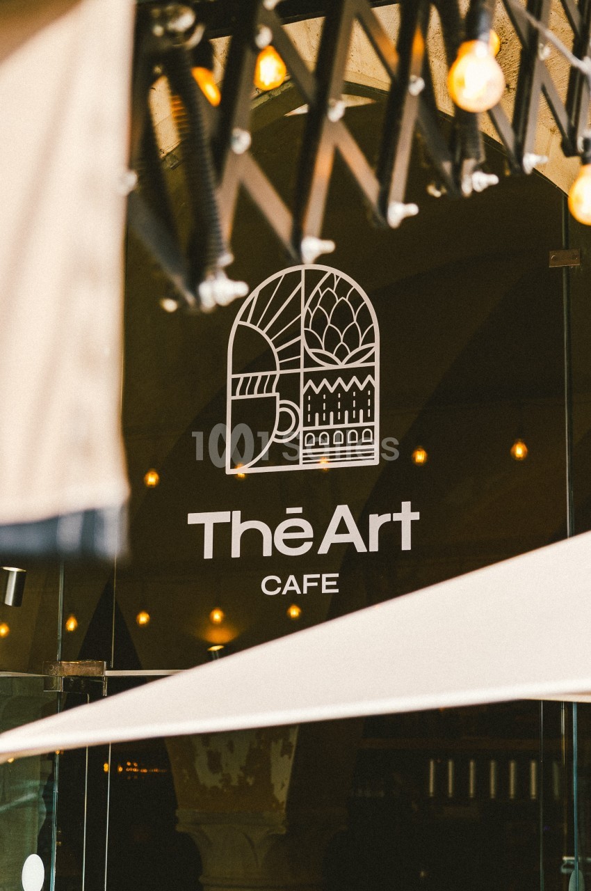 Logo ’The Art Café’ sur une vitre, entouré de lumières suspendues et d'éléments d'ombrage.