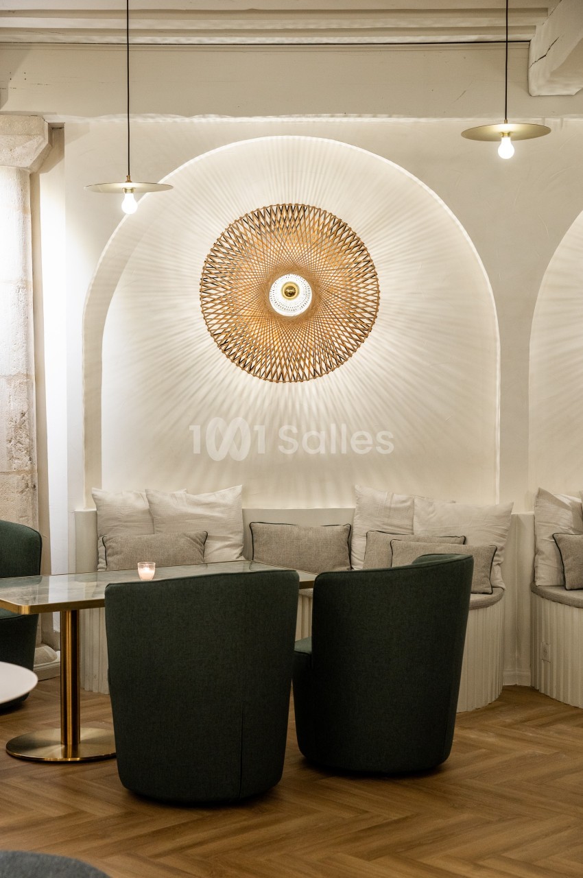 Espace intérieur avec fauteuils verts, table en verre, banquette beige et applique murale décorative en osier éclairée.
