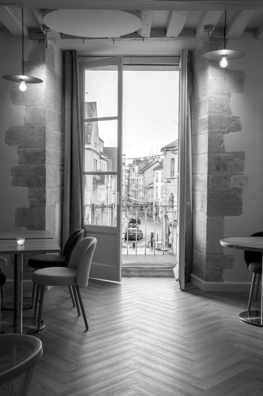 Salle lumineuse avec grandes fenêtres ouvertes sur une rue pavée, mobilier moderne et murs en pierre apparente.