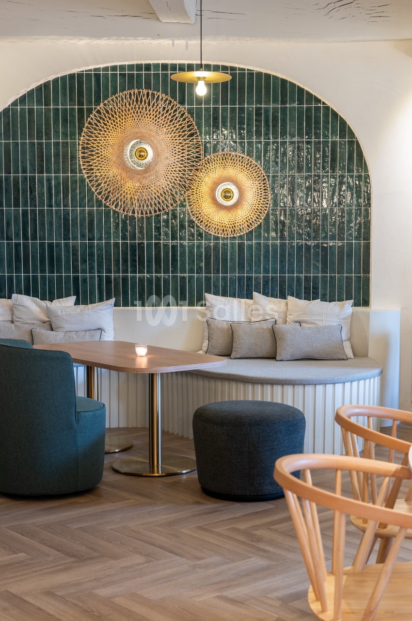 Coin salon avec table ronde, banquette, fauteuils et mur décoré de carreaux verts et luminaires dorés.