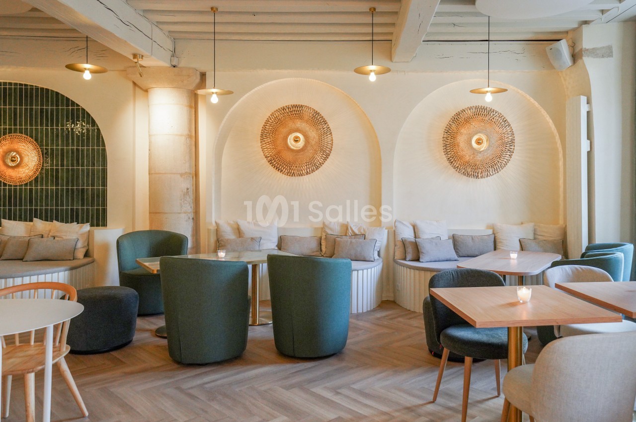 Salle lumineuse avec décoration murale en rotin, fauteuils verts, tables en bois et parquet clair.