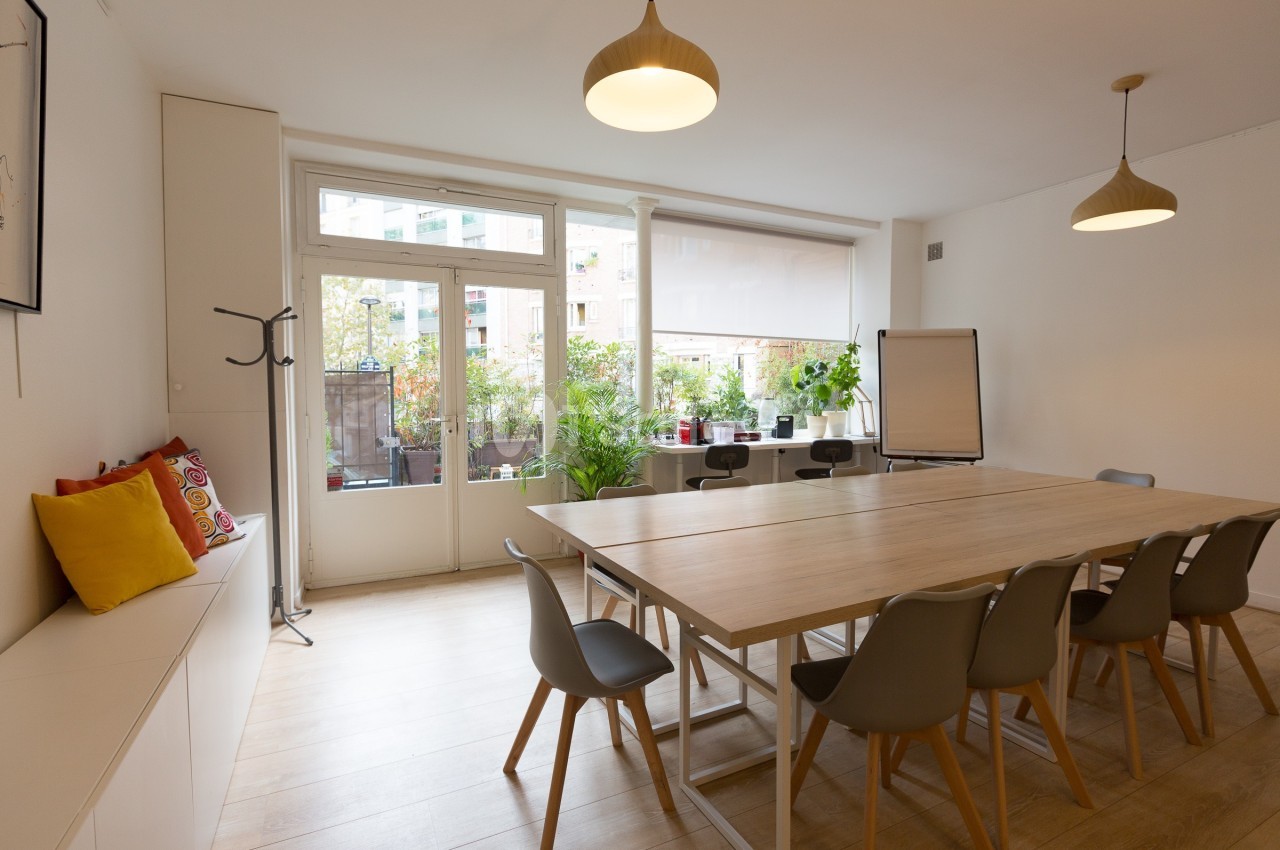 Salle de réunion lumineuse avec grande table en bois, chaises grises, tableau blanc et vue sur une terrasse végétalisée.