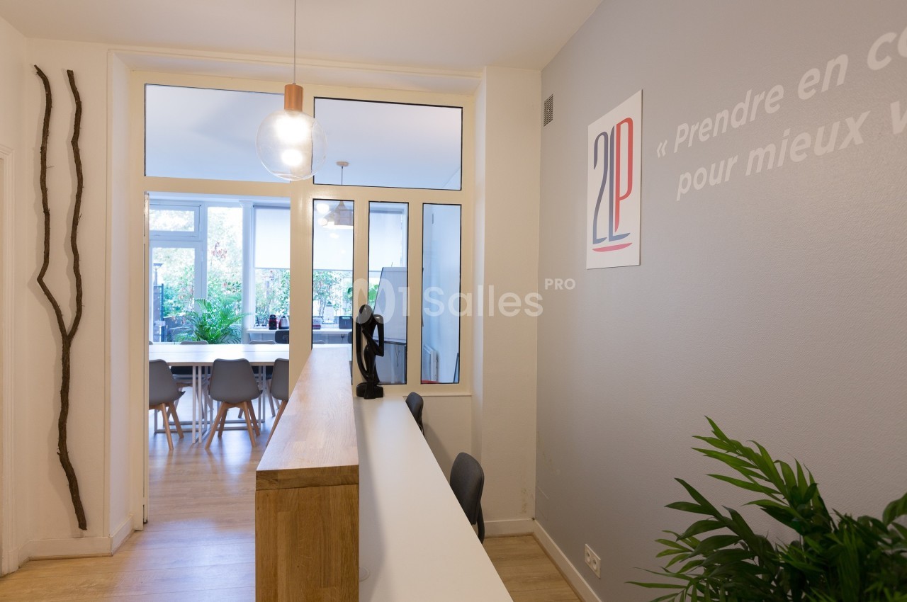 Bureau moderne avec comptoir d'accueil, plantes vertes et salle de réunion lumineuse en arrière-plan.