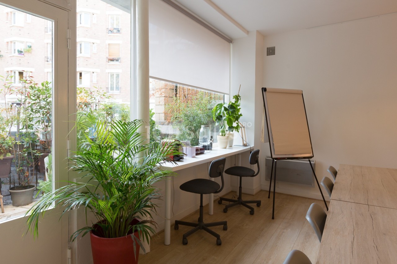 Salle lumineuse avec une grande fenêtre, des plantes, un bureau avec chaises, et un tableau de conférence.
