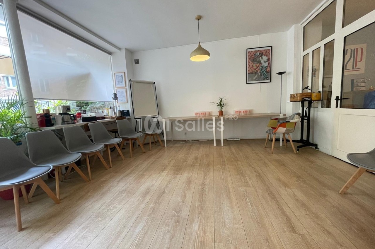 Salle lumineuse avec parquet, chaises disposées en cercle, tableau blanc et table avec plantes en arrière-plan.