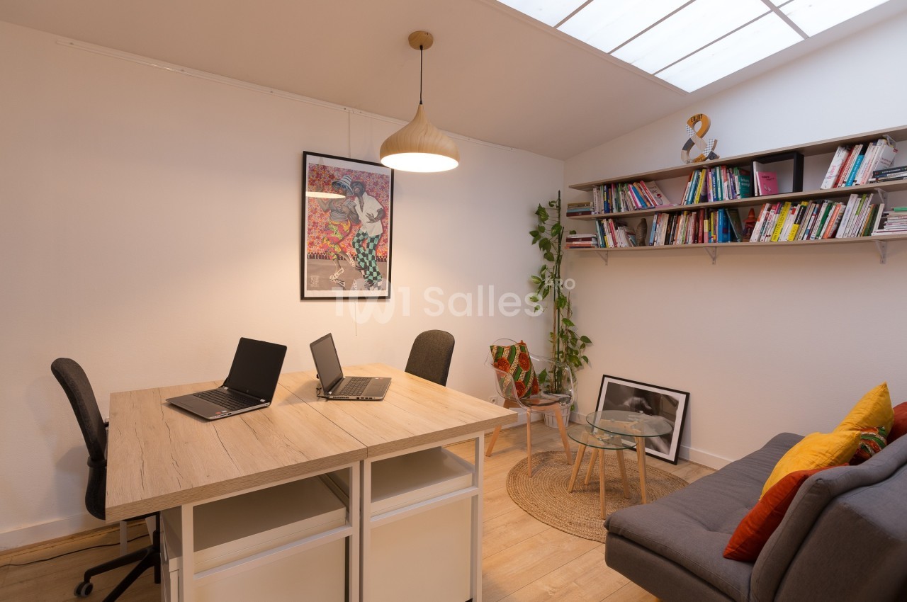 Bureau lumineux avec deux postes de travail, étagères remplies de livres, canapé et plantes décoratives.