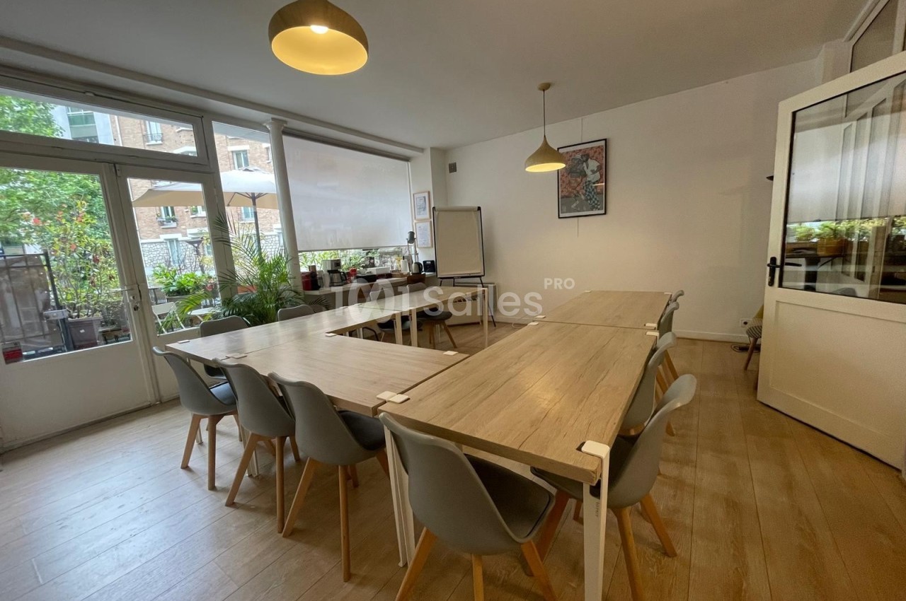 Salle lumineuse avec tables en bois, chaises grises, tableau blanc et vue sur une terrasse végétalisée.