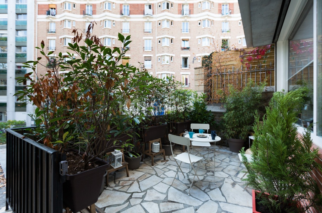 Terrasse en pierre avec mobilier de jardin, entourée de plantes en pots, donnant sur des immeubles en briques.