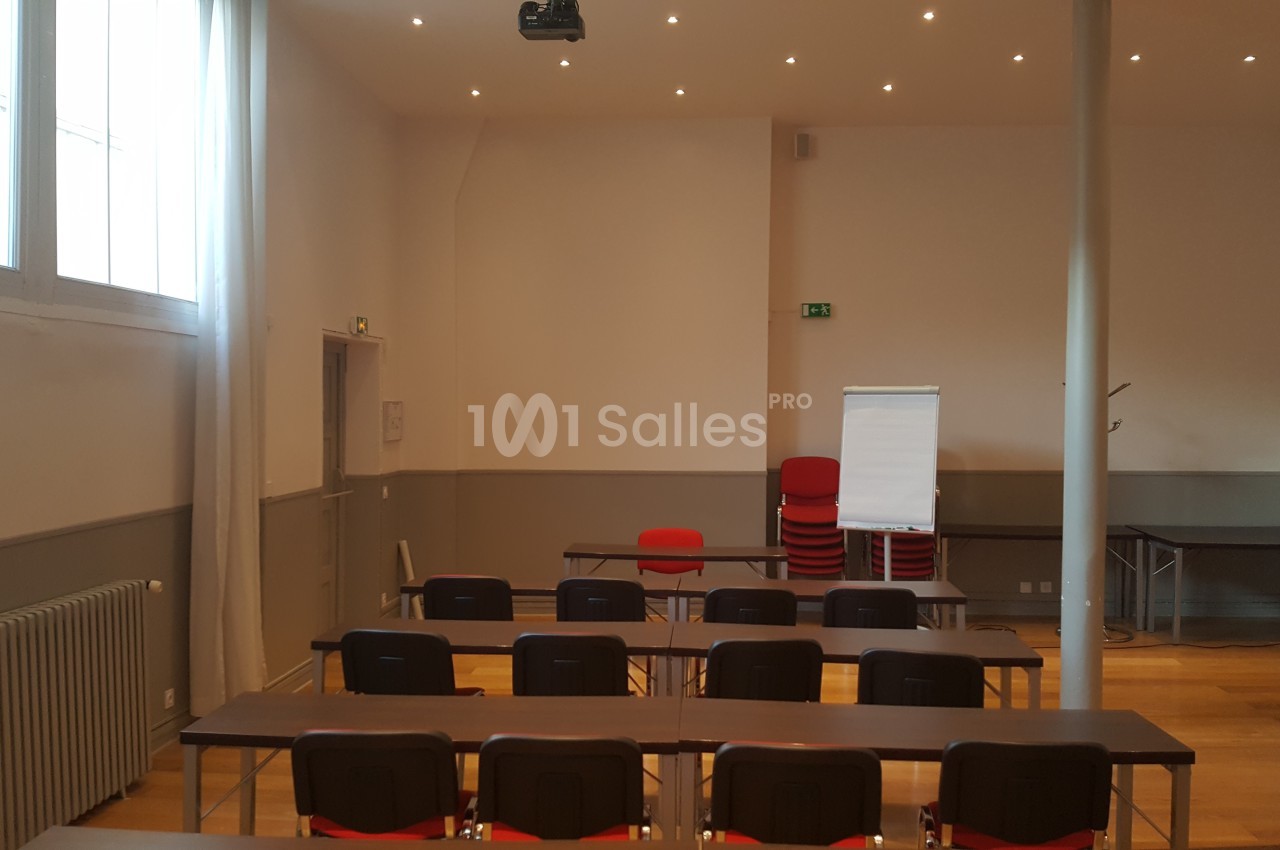 Salle de réunion lumineuse avec des tables, des chaises alignées, un paperboard et un projecteur au plafond.